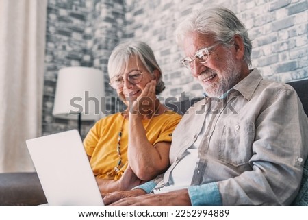 Pretty elderly 70s grey-haired couple resting on couch in living room hold on lap laptop watching movie smiling enjoy free time, older generation and modern wireless technology advanced users concept