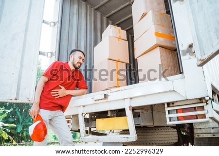 Delivery man having stomach ache while working with cardboard boxes behind container truck as background
