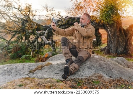 bearded man in speshial boots reaching the destination and resting under tree and taking photos on phone in Peak District at sunset on autumn day Travel Lifestyle concept