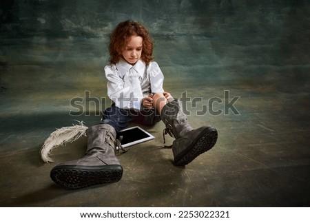 Wide angle view of little cute kid with curly hair sitting on floor over dark green background. Concept of childhood, comparison of eras, retro, vintage, emotions and kids fashion