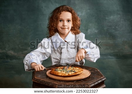 Tasting pizza. Portrait of little smiling girl with curly hair in vintage blouse eating pizza over dark green background. Concept of fast food, comparison of eras, retro, emotions. Looks happy