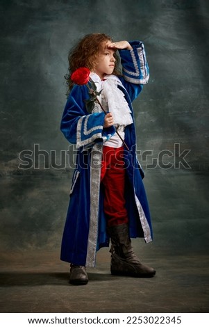 Emotional kid, little girl wearing costume of prince, musketeer and royal person posing over dark vintage style background. Fashion, theater, beauty, emotions concept. Eras comparison Royalty-Free Stock Photo #2253022345