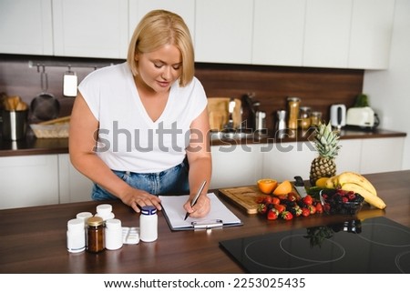 Caucasian young plump plus size woman counting calories, writing on clipboard healthy food, medicines, pills containers. Nutrition concept. Slimming shaping concept
