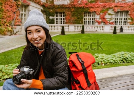 Beautiful young smiling asian woman in warm clothes taking pictures with vintage camera while sitting on bench in old city