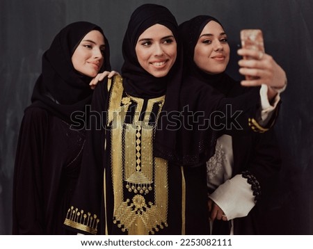 Group of young beautiful muslim women in fashionable dress with hijab using smartphone while taking selfie picture in front of black background