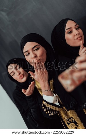 Group of young beautiful muslim women in fashionable dress with hijab using smartphone while taking selfie picture in front of black background