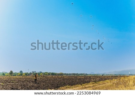 Back view of woman takes video clips and photo with tablet and watch with worry and concern during parachute training from airplane for army cadet in field with parachute and landscape in background.