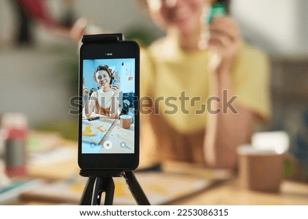 Focus on smartphone screen with happy young female artist showing paintbrush and small jar of gouache during online masterclass Royalty-Free Stock Photo #2253086315