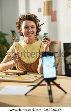 Happy young creative woman looking at smartphone camera during livestream or online masterclass for subscribers of her channel Royalty-Free Stock Photo #2253086317