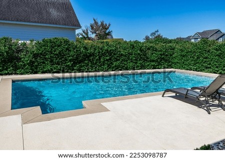 A rectangular new swimming pool with tan concrete edges in the fenced backyard of a new construction house.