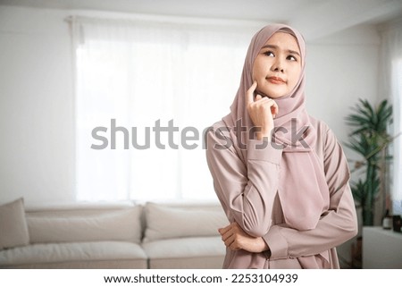 Beautiful smiling Asian woman in pink sweater keeps hands under chin, looks aside, remembers pleasant moment and positive thinking at modern home Royalty-Free Stock Photo #2253104939