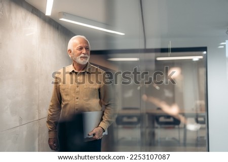 Smiling mature older successful gray-haired businessman leader, thoughtful senior professional businessman holding laptop looking away standing in modern office. Royalty-Free Stock Photo #2253107087