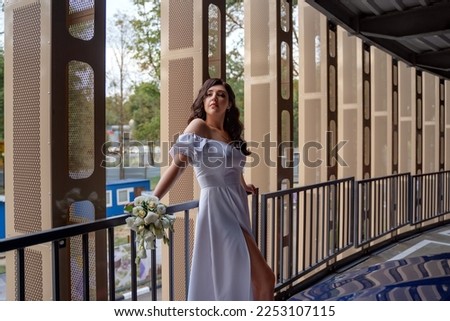 A young girl in a white dress stands at the railing at a height with a bouquet of flowers. Attractive brunette in the parking lot.