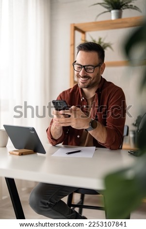 Happy smiling businessman wearing casual clothes and using modern smartphone in his home office, successful employer sitting at desk typing on mobile phone, also using digital tablet, pen and paper. Royalty-Free Stock Photo #2253107841