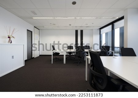 Spacious empty office with brown carpet and white wooden tables with swivel black seats and technical ceilings