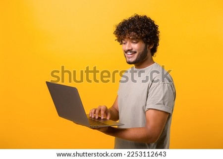 Smiling positive Indian man holding laptop in hands and typing. Blogger making posts in social networks, chatting with followers. Indoor studio shot isolated on yellow background