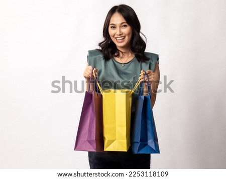 A portrait of a pretty Indonesian Asian girl in her 20s, wearing a light grey top and black trousers, looks excited while she opens up the shopping bag. Isolated on white background.