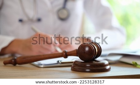 Wooden judge gavel on medical clinic desk and doctor doing paperwork in background. Legal support, malpractice and arbitration concept.