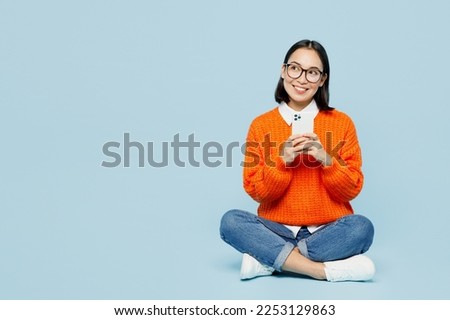 Full body young woman of Asian ethnicity wear orange sweater glasses hold in hand use mobile cell phone look aside on area isolated on plain pastel light blue cyan background. People lifestyle concept