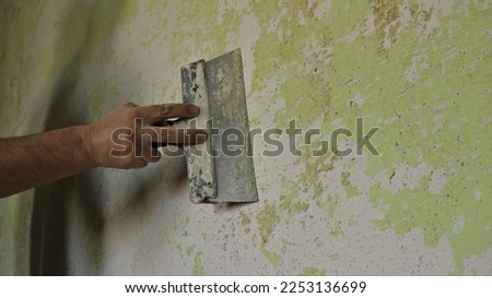 scraping off unevenness and residues of paint and plaster with a spatula, cleaning and preparing the surface of a room wall before sticking wallpaper, a hand scrapes off a layer on a dilapidated wall Royalty-Free Stock Photo #2253136699
