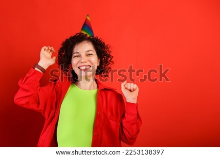 Curly cute girl laughs and jumps in a colorful birthday cap. Studio portrait of white woman on a bright red background. Positive smiling young female. Birthday greetings. Copy space. Royalty-Free Stock Photo #2253138397