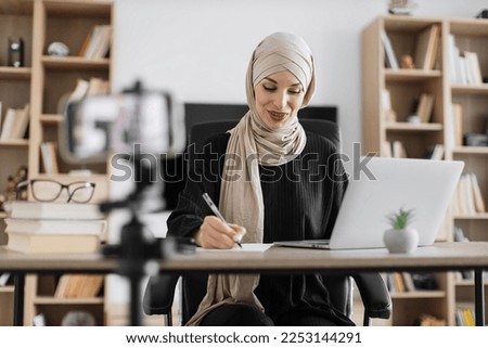 Cheerful young muslim woman in hijab recording creative video blog for her social networks, media for followers while working with laptop and writing report.