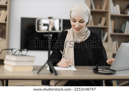 Charming muslim woman in hijab talking with subscribers while writing report and recording video on modern phone on selfie stick. Female blogger sitting at home and doing live stream.