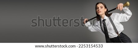 brunette woman in fashionable attire looking at camera while posing with walking stick on grey background, banner