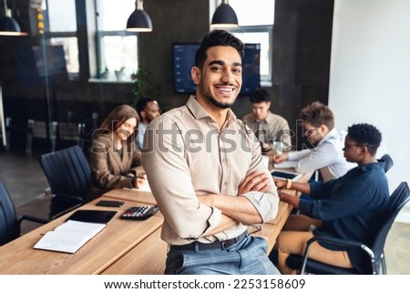 Successful person. Confident arab businessman leaning on desk in office, posing with folded arms and smiling at camera. Diverse colleagues working in background Royalty-Free Stock Photo #2253158609