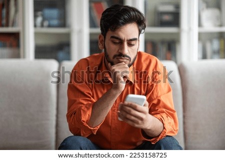 Thoughtful handsome young middle eastern man sitting on couch alone and using newest phone at home, concentrated arabic guy reading blog or news on Internet, touching his face, copy space Royalty-Free Stock Photo #2253158725