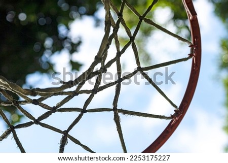 close-up of a basketball hoop with worn and dirty ropes and an orange hoop and in the background the blue sky and some blurred tree leaves.