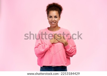 Confident African American woman in pink sweatshirt, holding hands at heart level, smiling looking at camera isolated on pink background. Breast Cancer Awareness Day. Cancer Campaign. Pink October 1st Royalty-Free Stock Photo #2253184429