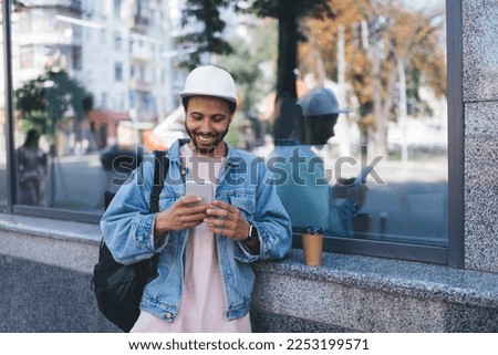 Millennial Hispanic tourist smiling in city using cellular application for tracking location GPS during travel vacations, cheerful hipster guy in white cap enjoying online networking and chatting