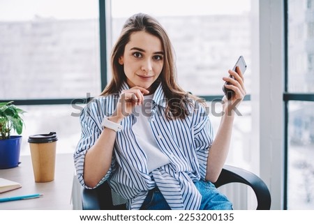 Portrait of Caucasian female employee in smart casual outfit sitting at table desktop with disposable takeaway cup and looking at camera during job break for social networking, wireless mobility Royalty-Free Stock Photo #2253200135
