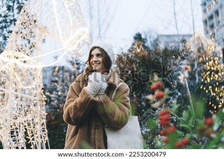 Happy young female in warm outerwear with shopper bag walking in Christmas market with garland lights and decorated trees behind