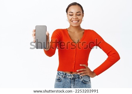 Portrait of 30 years old woman showing smartphone screen, demonstrates app interface, webstore, stands with mobile phone against white background.
