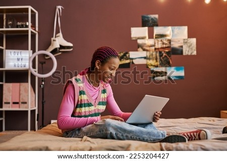 Side view portrait of black teenage girl using laptop while sitting on bed at home and smiling Royalty-Free Stock Photo #2253204427