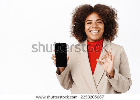 Smiling african american businesswoman promotes application, mobile app, shows telephone screen and okay sign, stands over white background.