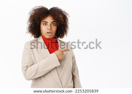 Shocked businesswoman points right direction and looks impressed, startled by smth, stands in suit over white background.