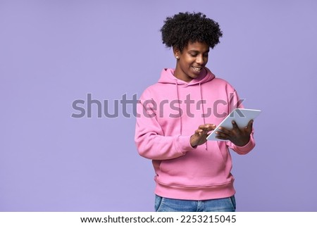 Young happy cool curly African American teenager student boy wearing pink hoodie holding pad using digital tablet computer technology browsing, elearning standing isolated on light purple background. Royalty-Free Stock Photo #2253215045