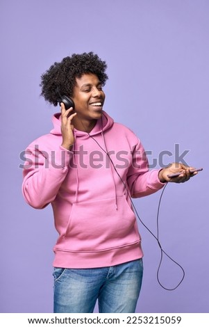 Happy cool African American teenager wearing headphones holding cell using smartphone dancing listening music on mobile phone technology, standing isolated on light purple background. Vertical