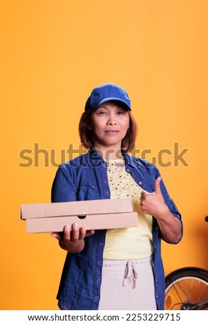 Asian pizzeria delivery employee doing thumbs up gesture while delivering carton flatbox with pizza to customers. Restaurant worker bringing orders with bike. Take out food service and concept