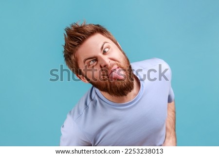 Portrait of crazy childish handsome young adult bearded man looking at camera with crossed eyes and showing tongue out, expressing positive emotions. Indoor studio shot isolated on blue background. Royalty-Free Stock Photo #2253238013