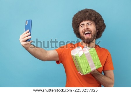 Portrait of happy joyful man with Afro hairstyle wearing orange T-shirt having video call with present box, boasting his gift to followers. Indoor studio shot isolated on blue background.
