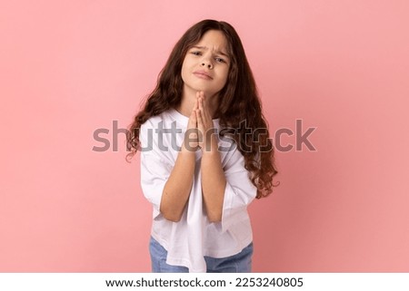 Please help me or forgive. Portrait of little girl wearing white T-shirt standing with palm hands and looking at camera pleased pleading. Indoor studio shot isolated on pink background. Royalty-Free Stock Photo #2253240805