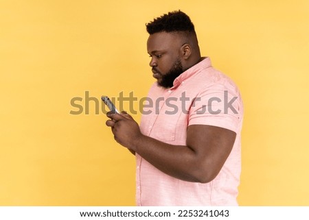 Side view of serious man wearing pink shirt sending text messages via mobile phone, searches gifts for holiday in internet, uses smartphone app. Indoor studio shot isolated on yellow background.