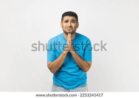 Portrait of sad upset unshaven man wearing blue T- shirt standing has sorrowful expression asking somebody to forgive, keeps palms in praying gesture. Indoor studio shot isolated on gray background. Royalty-Free Stock Photo #2253241417