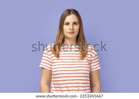 Portrait of serious confident blond woman wearing striped T-shirt standing looking at camera with attentive look, being strict and bossy. Indoor studio shot isolated on purple background. Royalty-Free Stock Photo #2253241667