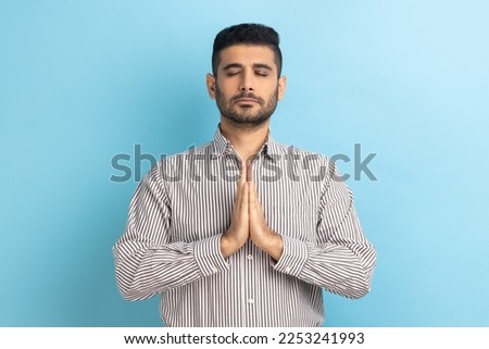 Portrait of calm relaxed young businessman with beard standing doing yoga meditating exercise, keeping palms together, wearing striped shirt. Indoor studio shot isolated on blue background.