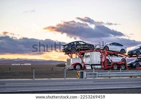 Industrial car hauler big rig white semi truck tractor transporting cars on the modular two level hydraulic semi trailer driving on the highway road between the field in California at twilight Royalty-Free Stock Photo #2253269459